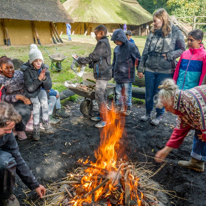 Eine Schulklasse sitzt um ein Feuer herum. Unter dem Feuer liegen die Steine zum Brotbacken. Das Feuer soll sie erhitzen. (vergrößerte Bildansicht wird geöffnet)