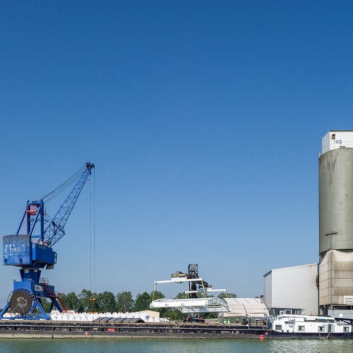 Eine Dachkonstruktion der Firma wird an einem Hafen auf ein Schiff geladen. Im Vordergrund das Schiff, dahinter ein großer blauer Kran. (vergrößerte Bildansicht wird geöffnet)